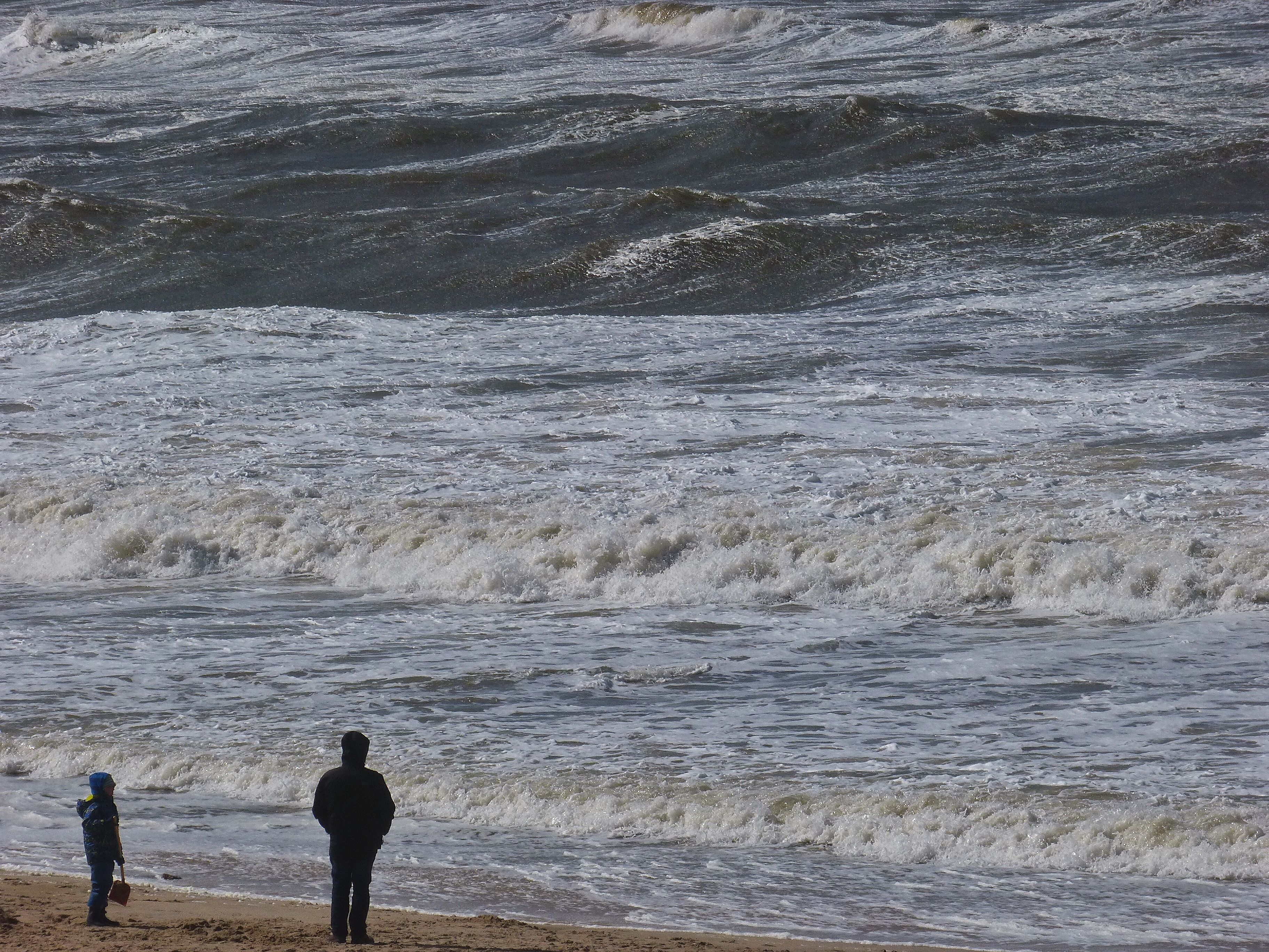 Starke Wellen schlagen an den Sylter Strand, am Strand stehen zwei Personen und schauen auf die Wellen hinaus