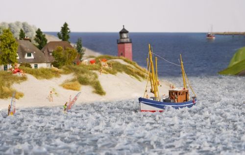 Ein Modell-Kutter fährt auf dem Wasser an einem kleinen Häuschen und einem Leuchtturm vorbei