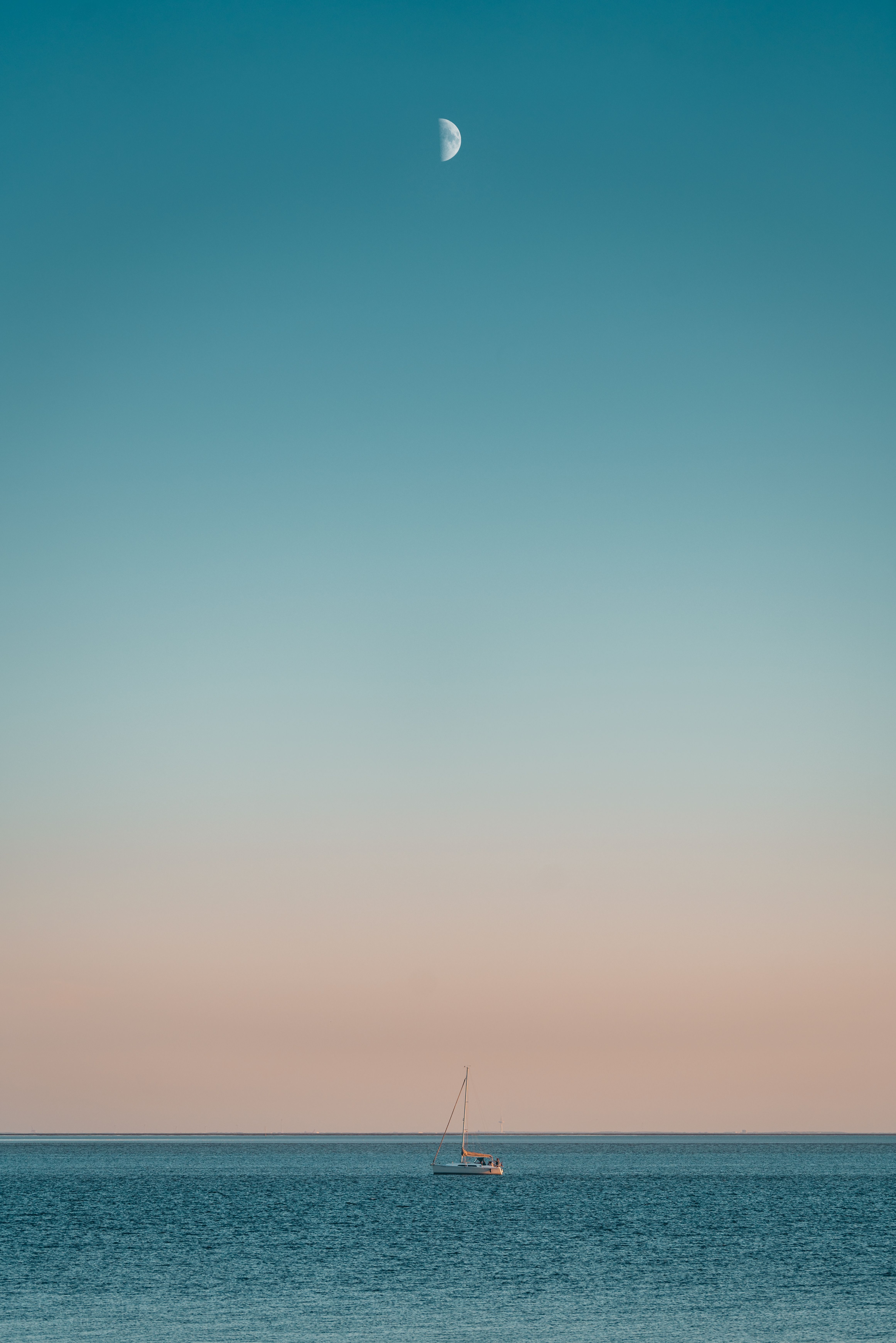 Ein Segelboot liegt vor der Küste von Büsum. Die Sonne geht gerade unter und über dem Boot steht bereits der Mond.