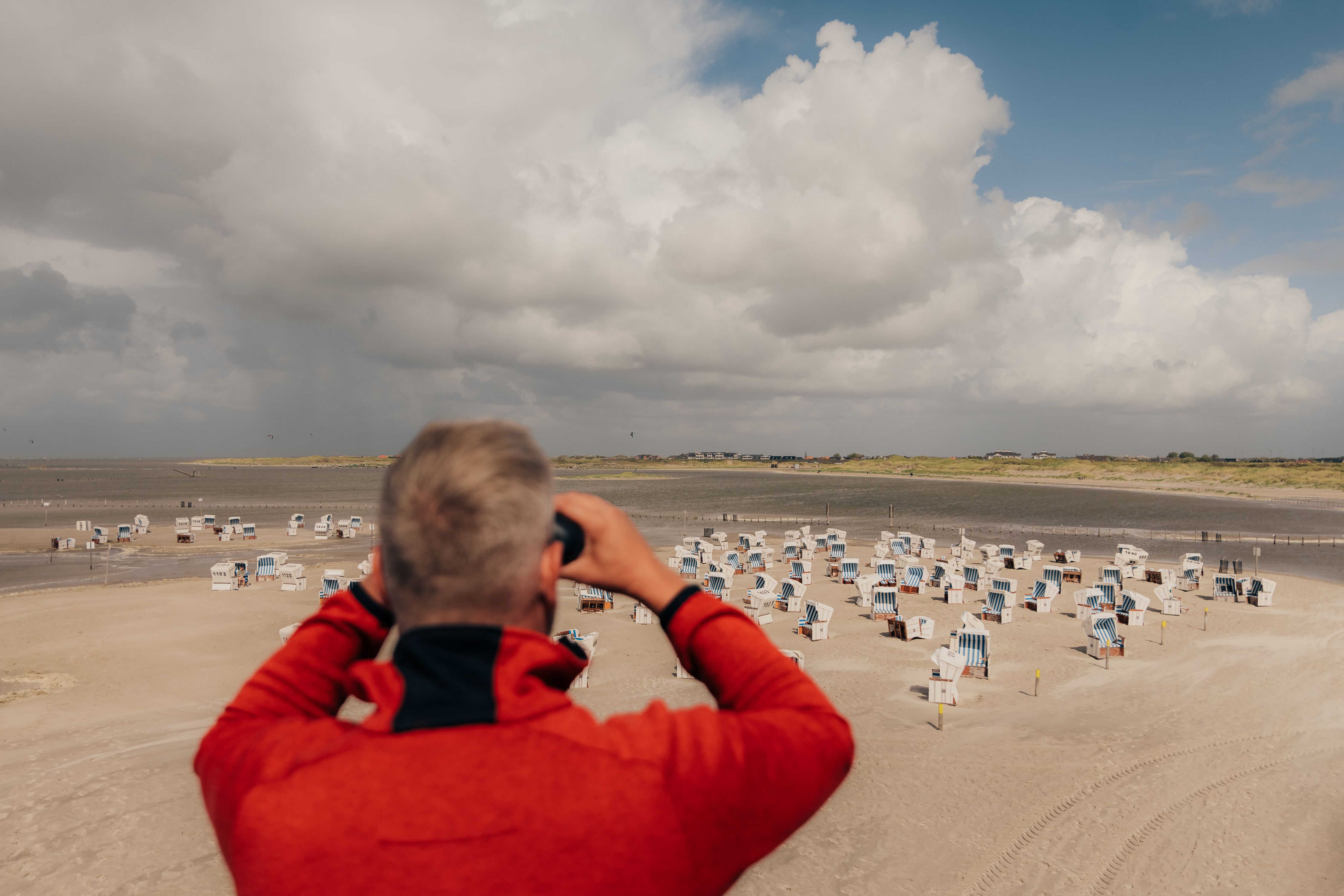 Ein Mann schaut mit einem Fernglas über den Strand von St. Peter-Ording, auf dem viele Strandkörbe stehen