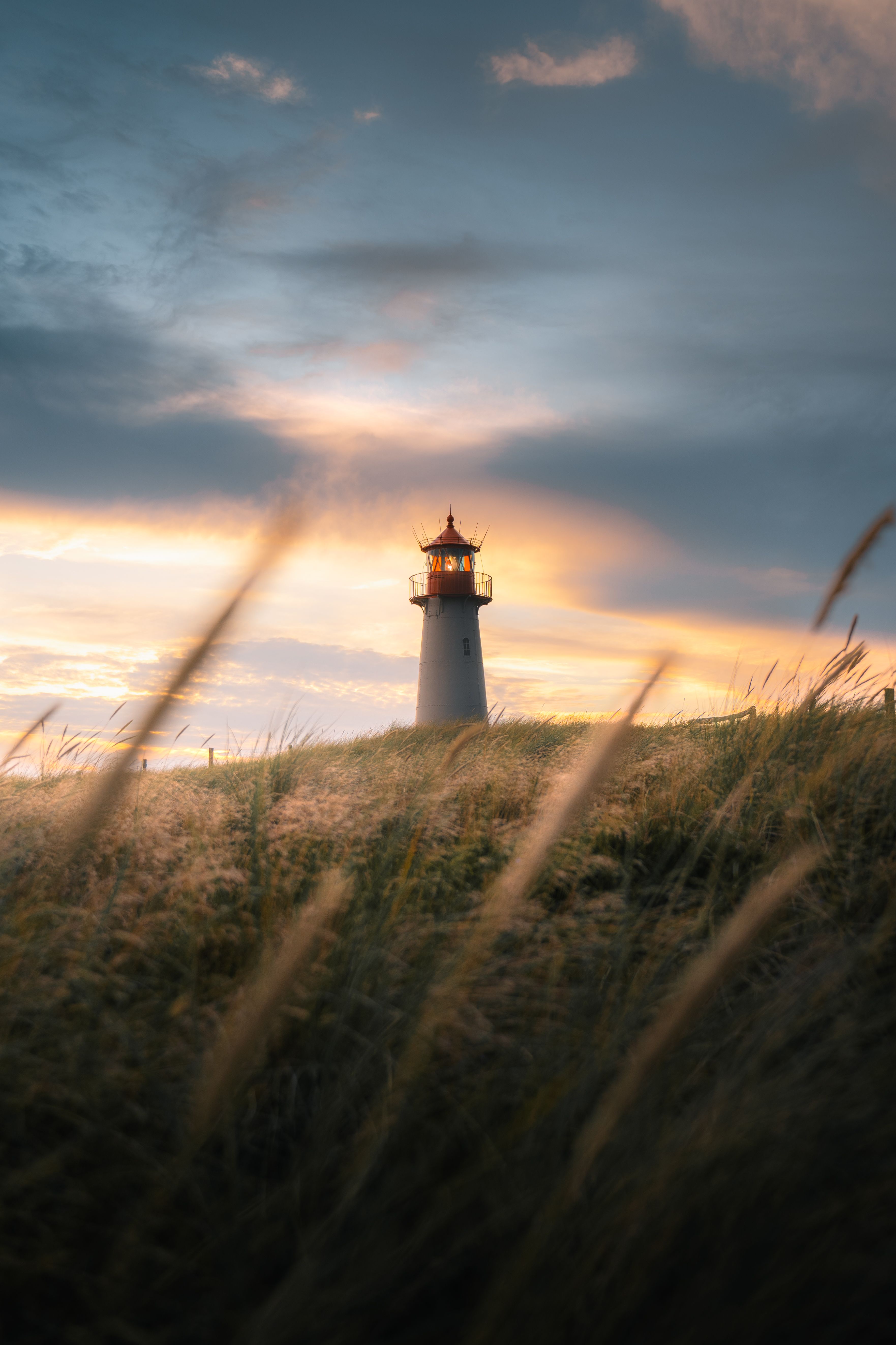 Blick auf einen Leuchtturm, im Vordergrund ist das Dünengras zu erkennen, hinter dem Leuchtturm bilden Wolken ein schönes Spektakel am Himmel