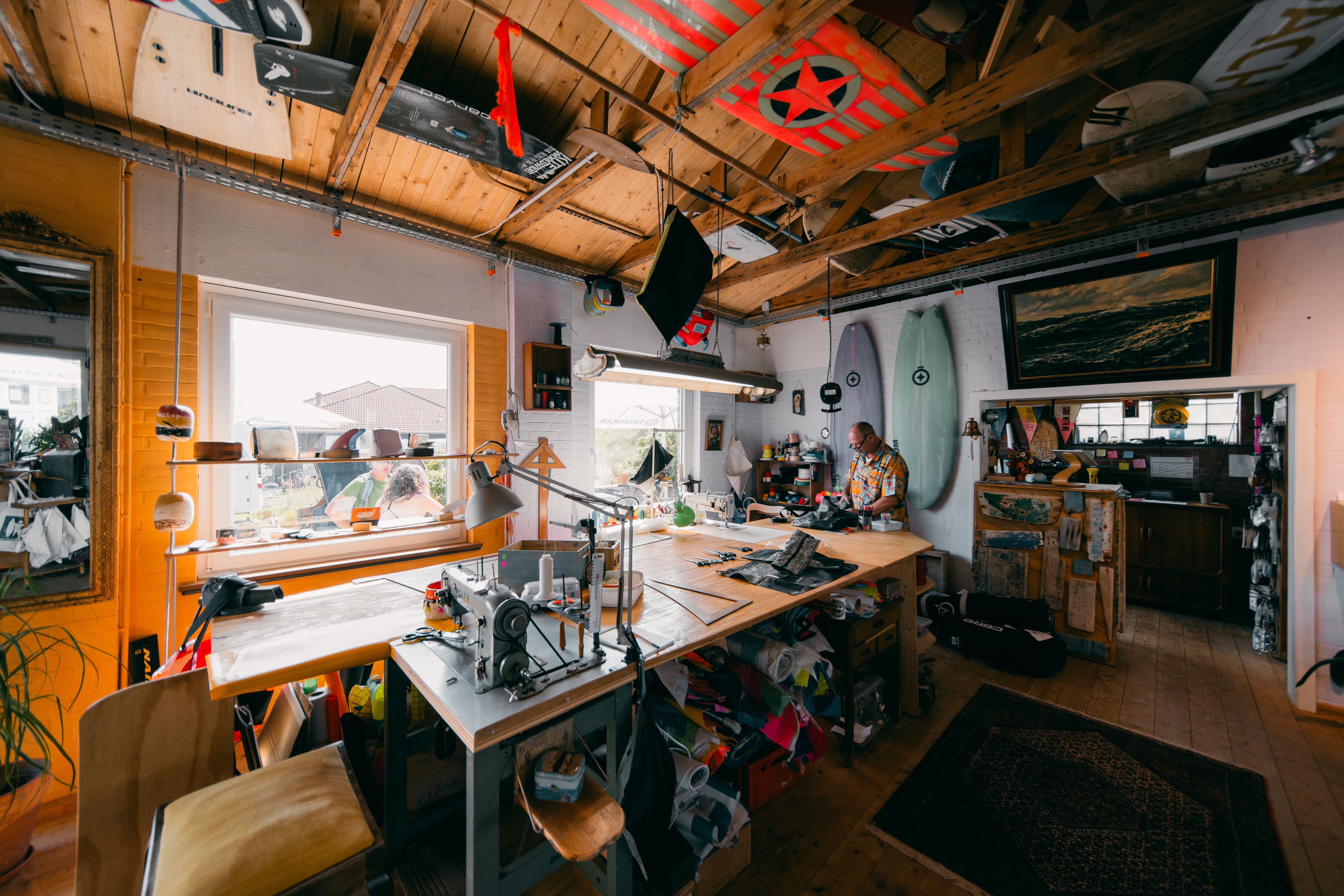 Weitwinkelaufnahme vom Surfhouse in Westerland auf Sylt. Mitarbeiter Andy sitzt an seiner Nähmaschine und näht Taschen aus alten Surfmaterialien und Strandkorbinnenleben.