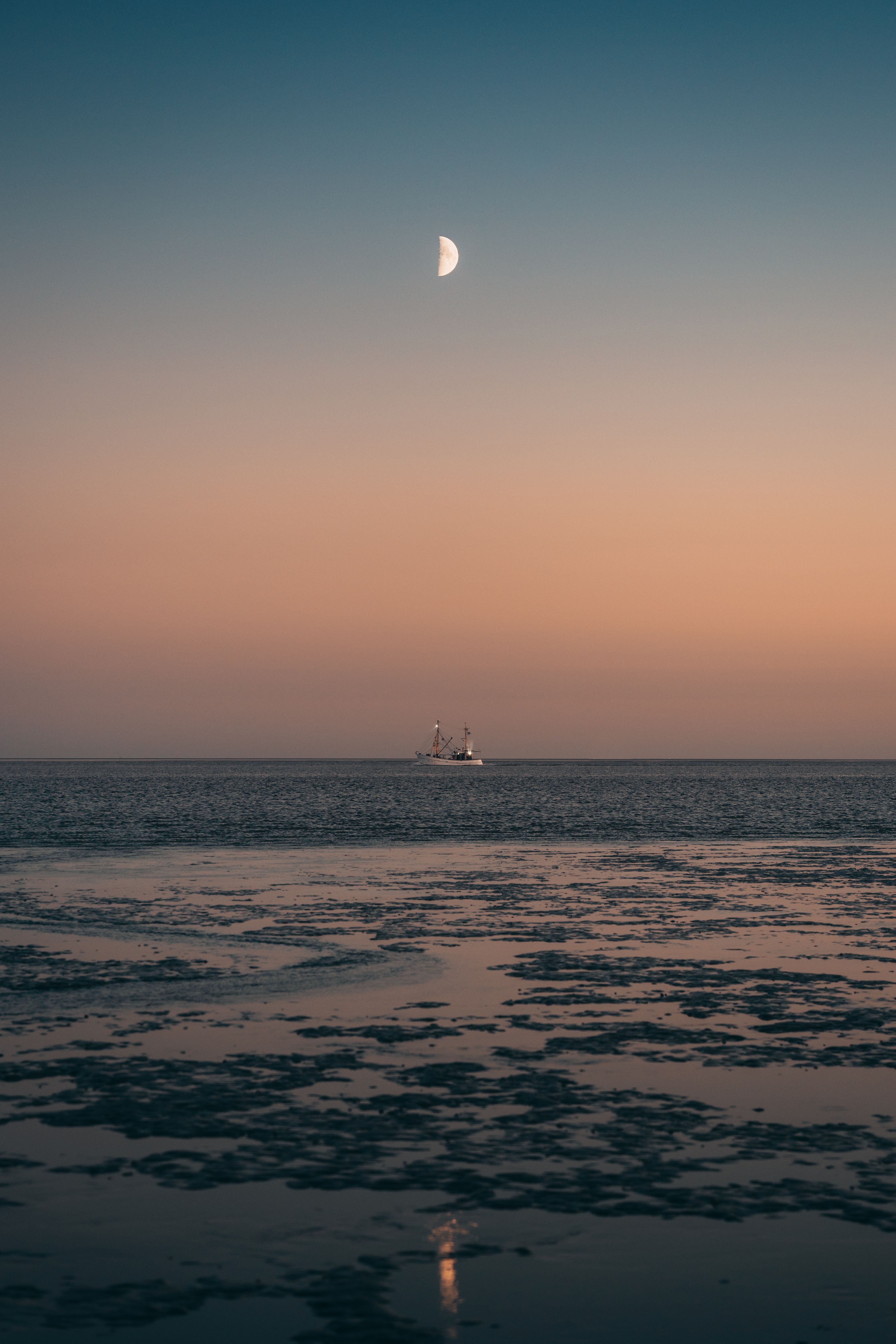 Blick über das Wattenmeer hinzu einem Krabbenkutter, über ihm ist der Halbmond an einem rötlichen Himmel zu sehen