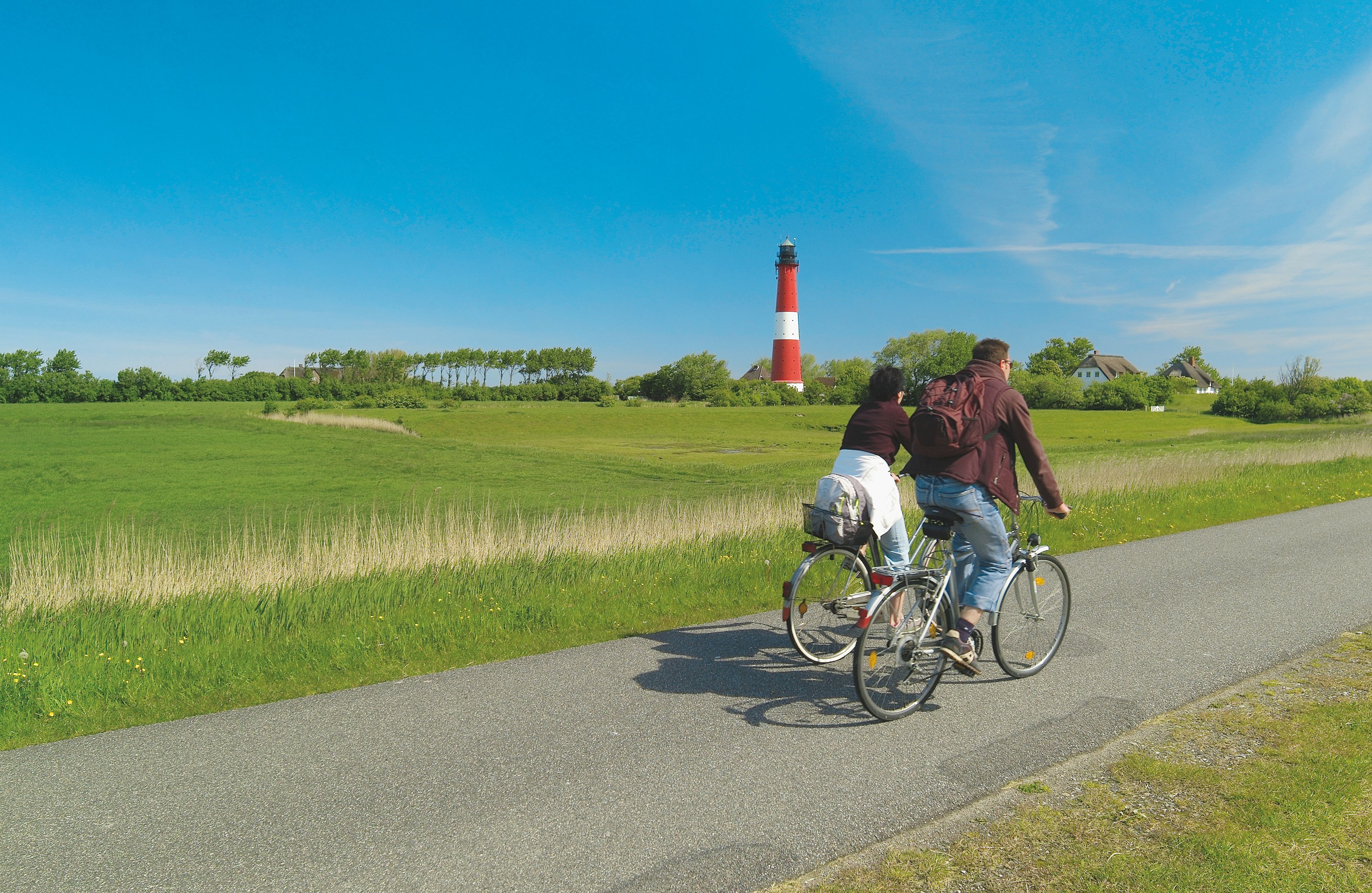 Zwei Personen fahren hinter dem Deich entlang, im Hintergrund ist der Pellwormer Leuchtturm zu sehen