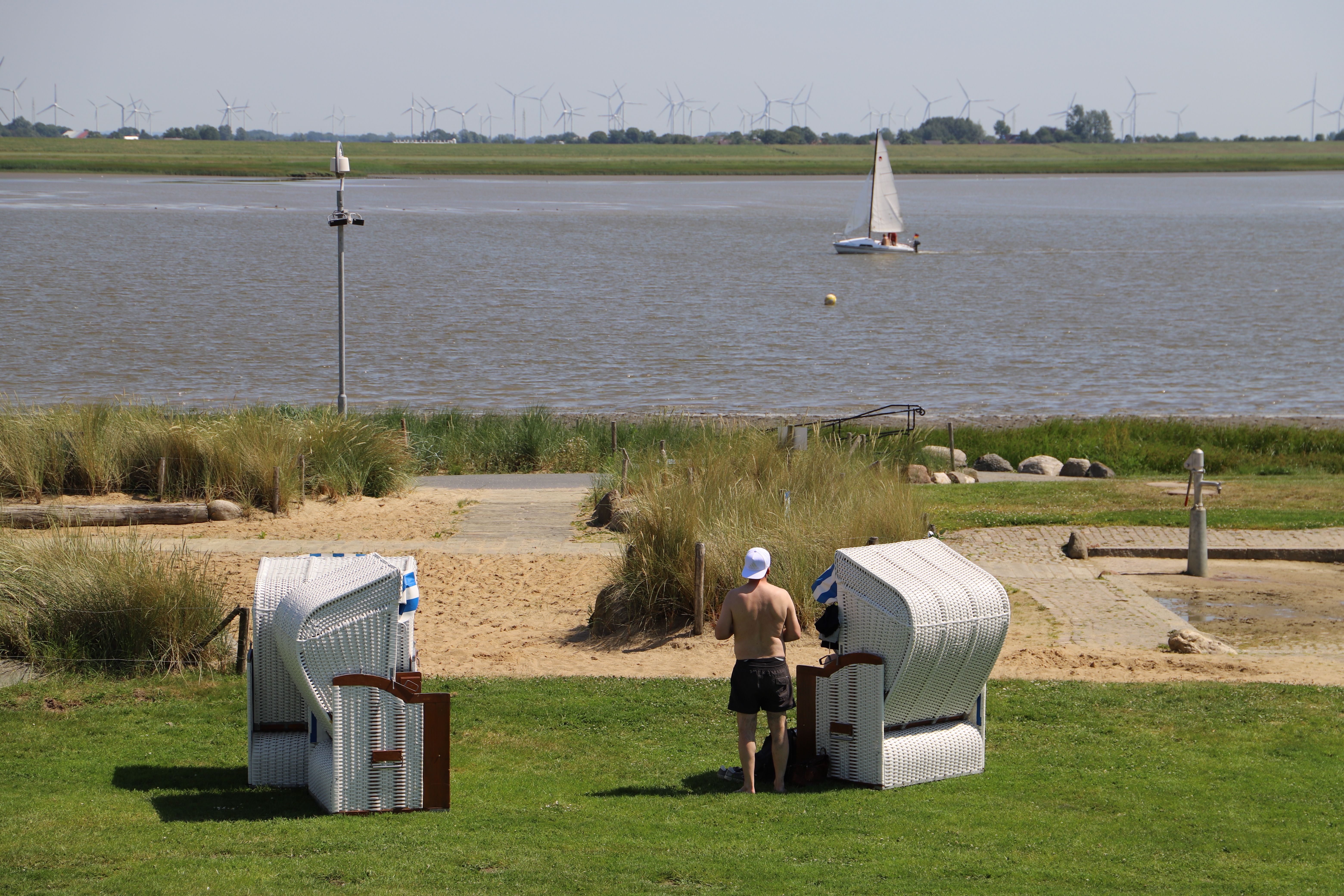 Zwei Strandkörbe stehen auf dem Deich und eine Person blickt zu einem Segelboot, das auf dem Meer entlangfährt