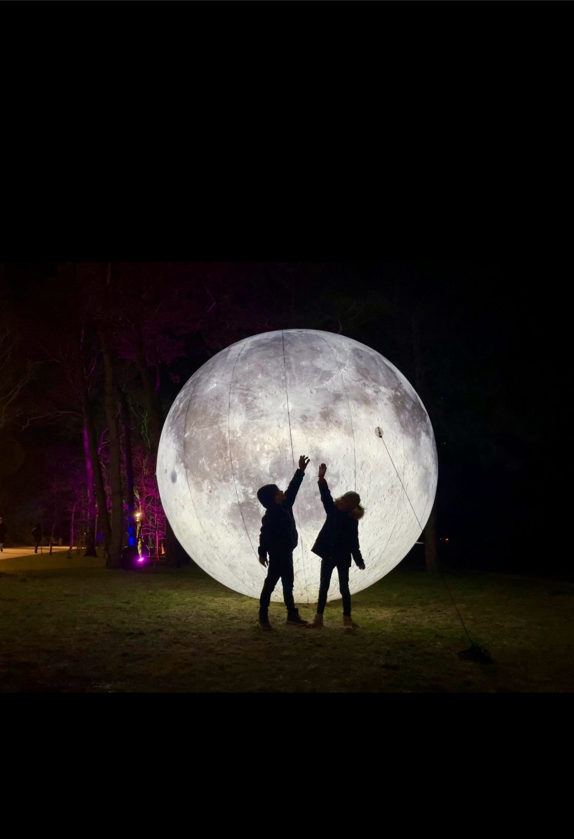 Zwei Kinder stehen vor einem großen, aufblasbaren Mond und strecken je eine Hand nach oben